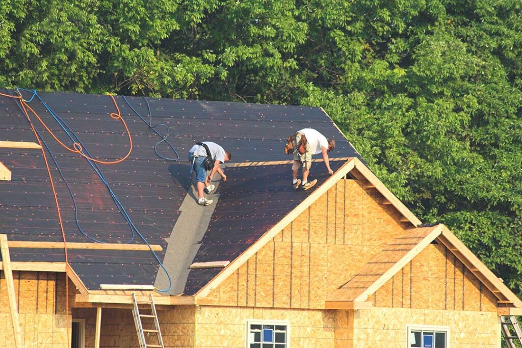 A person safely clearing leaves and debris from a house roof using a ladder and gloves on an overcast autumn day.
