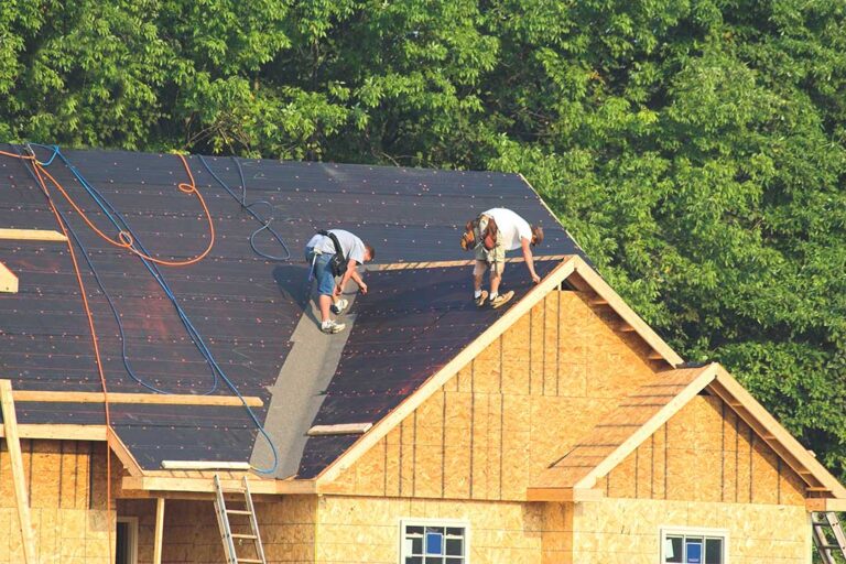 A person safely clearing leaves and debris from a house roof using a ladder and gloves on an overcast autumn day.