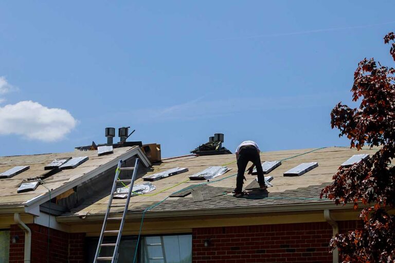 Person inspecting damaged shingles and water stains on a house roof with visible wear and missing sections.