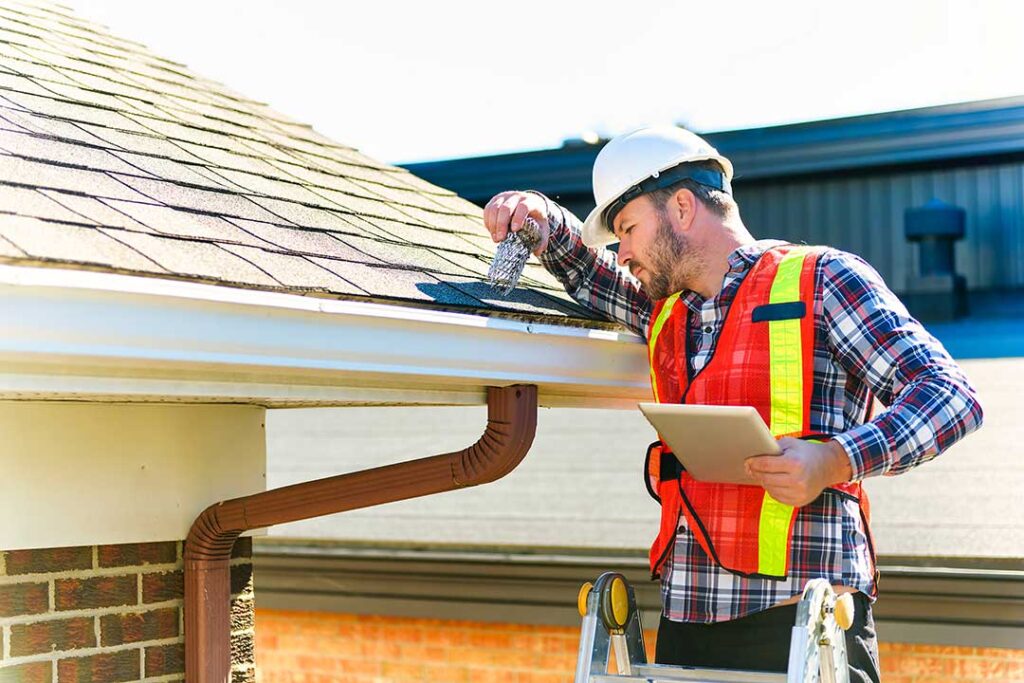 Roofing worker rolls underlayment across a wood roof deck beside stacked shingles.
