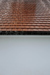 Snow rests on a residential shingle roof while a person examines the eaves and gutters from the ground.