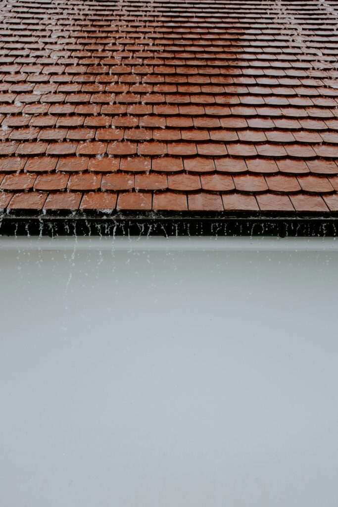 Snow rests on a residential shingle roof while a person examines the eaves and gutters from the ground.